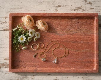 May include: A rectangular red stone tray holding a small bouquet of pale peach ranunculus and white daisies, two gold rings, and a gold chain necklace with a clear, teardrop-shaped pendant resting on the textured, porous surface.