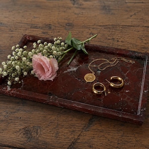 Könnte beinhalten: Ein rechteckiges Tablett aus dunkelrotem Marmor mit polierter Oberfläche. Auf dem Tablett liegen eine goldfarbene Halskette mit rundem Anhänger, ein Paar goldfarbene Creolen, eine hellrosa Blume und ein Zweig mit weißem Schleierkraut.