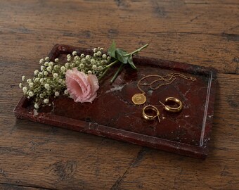May include: A rectangular dark red marble tray with a polished finish. Resting on the tray are a gold-toned necklace with a circular pendant, a pair of gold-toned hoop earrings, a single light pink flower, and a sprig of white baby's breath.