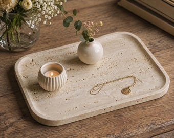 May include: A rectangular beige travertine stone tray with a porous, textured surface. On the tray sits a small, round, off-white ceramic vase containing a single sprig of greenery, a ribbed white candle holder with a lit candle, and a gold necklace.