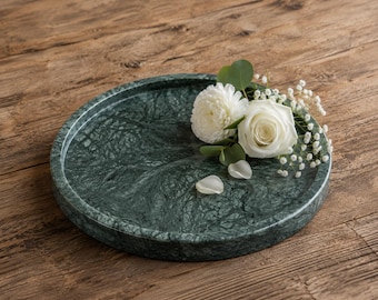 May include: A circular dark green marble tray with a polished finish and natural veining patterns. The tray is adorned with a small arrangement of white flowers, including a rose and a carnation, accented by green eucalyptus leaves and baby's breath.