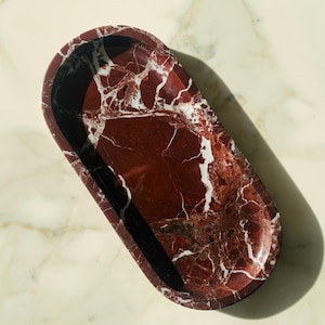 Puede incluir: Bandeja ovalada con un llamativo patrón de mármol rojo y vetas blancas. El interior de la bandeja es rojo oscuro con bordes negros, creando un contraste. Mide aproximadamente 30 cm de largo y 15 cm de ancho, ideal para decoración.
