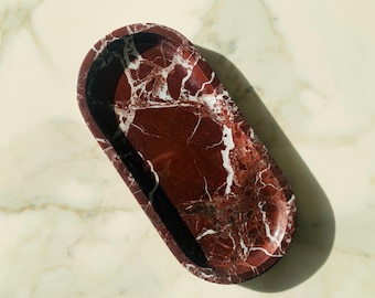May include: Oval-shaped tray with a striking red marble pattern, featuring white veining. The tray's interior is a deep red with black edges, creating a contrasting border. The tray is approximately 12 inches long and 6 inches wide.