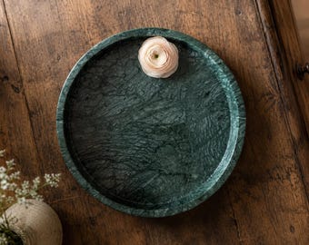 May include: A circular dark green marble tray with a polished finish and natural veining patterns. A single pale pink ranunculus flower rests near the top edge of the tray, which features a raised rim and a smooth, flat interior surface.