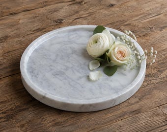 May include: A circular white marble tray with grey veining, featuring a small arrangement of white ranunculus, a pale pink rose, green eucalyptus leaves, and delicate white baby's breath flowers resting on the surface of the stone.