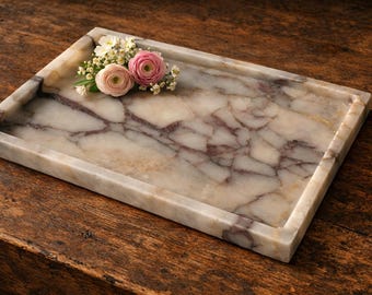May include: A rectangular marble tray with a cream and burgundy veined pattern. The tray has a raised edge and is decorated with a small arrangement of pink and white flowers. The tray is displayed on a dark wood surface.