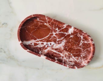 May include: A red marble oval tray with white veining. The tray has a smooth, polished surface and a shallow depth. The tray is a decorative item that can be used to hold jewelry, keys, or other small items. The tray is approximately 10 inches long.
