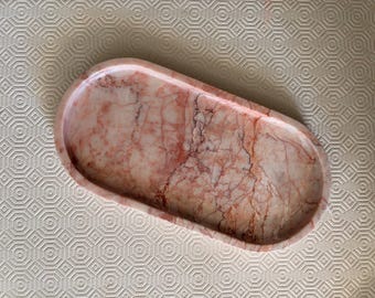 May include: An oval-shaped tray crafted from polished stone featuring a natural marble pattern with swirling veins of soft pink, peach, and deep reddish-brown hues. The tray has a smooth, concave surface with raised edges and a glossy finish.