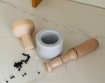 May include: A mortar and pestle set with a white marble bowl and wooden pestle. The set includes a separate wooden pestle and a small pile of black peppercorns. The items are arranged on a white marble surface.
