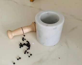 May include: A white marble mortar and pestle set with a light wood pestle. Black peppercorns are scattered on the surface next to the mortar. The mortar is cylindrical and has a smooth, polished finish.