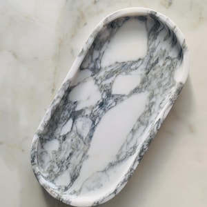 Könnte beinhalten: Ovale Marmorschale mit weißer Basis und grauer Maserung. Die Schale hat eine glatte, polierte Oberfläche und einen erhöhten Rand. Die Schale ist etwa 25 cm lang und 13 cm breit, ideal für die Präsentation von Schmuck oder kleinen Gegenständen.
