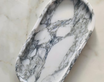May include: Oval marble tray with a white base and gray veining. The tray has a smooth, polished surface and a raised edge. The tray is approximately 10 inches long and 5 inches wide, perfect for displaying jewelry or small items.