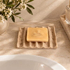 May include: A rectangular beige stone soap dish with a wavy, grooved surface texture. A single, solid, light yellow rectangular bar of soap rests centered on the dish. The soap dish features a natural, speckled, porous appearance with visible indentations.