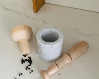 May include: A mortar and pestle set with a white marble mortar and a wooden pestle. The set is accompanied by a wooden bowl and scattered peppercorns, ideal for grinding spices. The items are arranged on a white surface.