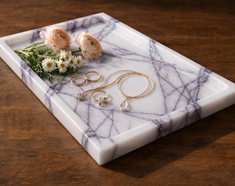 May include: A rectangular white marble tray with purple veining. The tray holds two pale pink ranunculus flowers, a small cluster of white daisies, two gold rings, and a gold necklace featuring a clear teardrop-shaped pendant.