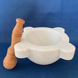 May include: A white marble mortar and pestle set. The mortar has a round bowl with four flared edges. The pestle is made of light-colored wood with a rounded head and a handle with carved rings.