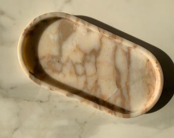 May include: Oval-shaped decorative tray with a marble design. The tray has a light beige background with brown veining throughout. The tray is smooth and has rounded edges. The tray is sitting on a marble surface.