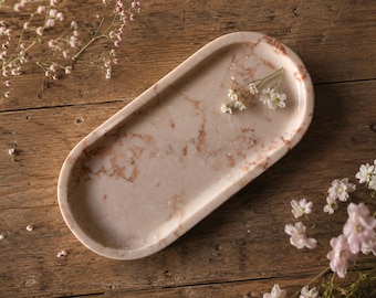 May include: An oval-shaped tray crafted from polished stone featuring a light beige base with prominent reddish-brown veining throughout. The tray has a smooth, raised rim and a shallow basin, displaying a natural marbled pattern across its surface.