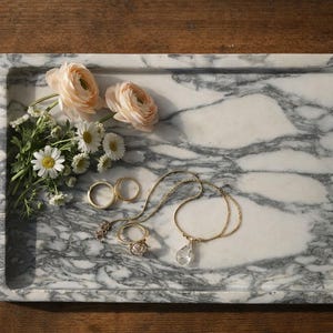 May include: A marble tray with a gray and white pattern holds a collection of jewelry and flowers. Two gold rings, a necklace with a clear pendant, and a ring with a cluster of stones are displayed. Soft pink flowers and white daisies add a delicate touch.