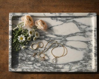 May include: A marble tray with a gray and white pattern holds a collection of jewelry and flowers. Two gold rings, a necklace with a clear pendant, and a ring with a cluster of stones are displayed. Soft pink flowers and white daisies add a delicate touch.