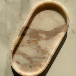 Könnte beinhalten: Eine ovale Marmorschale mit einem natürlichen, geäderten Muster in Creme- und Pfirsichtönen. Die Schale hat eine glatte, polierte Oberfläche und sanft geschwungene Kanten. Diese dekorative Schale eignet sich für Schmuck oder kleine Gegenstände.