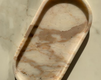 May include: An oval-shaped marble tray with a natural, veined pattern in shades of cream and peach. The tray has a smooth, polished surface and gently curved edges. This decorative tray is suitable for jewelry or small items.