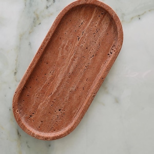 Può includere: Un vassoio di forma ovale realizzato in pietra di travertino color bruno-rossastro. La superficie presenta una consistenza naturale e porosa con striature lineari visibili e piccole rientranze caratteristiche della composizione organica.