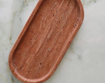May include: An oval-shaped tray crafted from reddish-brown travertine stone. The surface features a natural, porous texture with visible linear striations and small indentations characteristic of the stone's organic composition and raw finish.