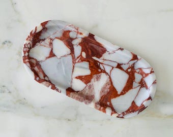 May include: Oval marble tray with a striking pattern of white and reddish-brown veining. The tray has a smooth, polished surface and a slightly raised edge. The tray is designed to hold small items.
