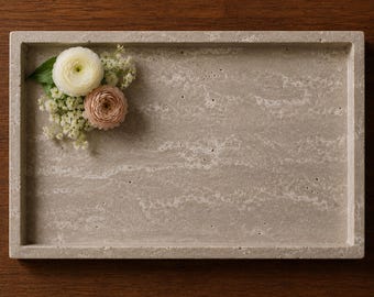 May include: A rectangular beige stone tray with a natural porous texture and visible veining. A small cluster of white and pale pink ranunculus flowers with green leaves and tiny white filler blossoms is placed in the upper left corner.