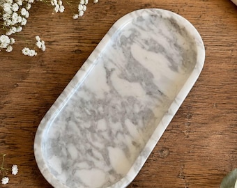 May include: An oval-shaped tray crafted from white marble with prominent grey veining throughout the surface. The tray features a smooth, polished finish and a raised rim, showcasing the natural stone pattern with its unique swirls and grey color variations.