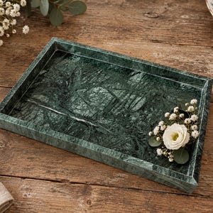Può includere: Un vassoio rettangolare realizzato in marmo verde scuro con venature bianche prominenti. Il vassoio presenta bordi rialzati e una superficie liscia e lucida. Un piccolo mazzo di fiori bianchi e foglie verdi riposa all'interno sul lato destro.