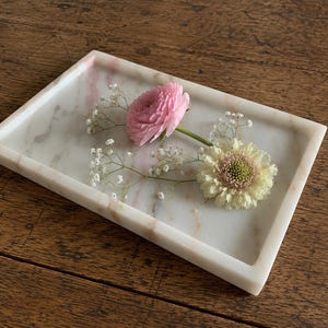 Op de afbeelding: Een rechthoekig dienblad van wit marmer met subtiele roze en grijze aders. In het dienblad liggen twee verse bloemen, een roze ranonkel en een crèmekleurige scabiosa, vergezeld door kleine takjes witte gipskruidbloemen.