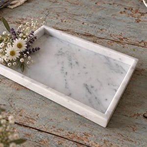 May include: A rectangular white marble tray with grey veining and raised edges. A small decorative arrangement of white daisies, purple lavender sprigs, and baby's breath flowers is placed on the top left corner of the tray surface.