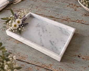 May include: A rectangular white marble tray with grey veining and raised edges. A small decorative arrangement of white daisies, purple lavender sprigs, and baby's breath flowers is placed on the top left corner of the tray surface.
