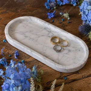 Op de afbeelding: Een ovaal wit marmeren dienblad met grijze aders waarop drie ringen liggen. Het dienblad heeft een gladde, gepolijste afwerking en een opstaande rand, wat een strakke presentatieruimte biedt voor de twee goudkleurige banden en één zilverkleurige ring.