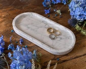 May include: An oval white marble tray with grey veining featuring three rings resting on its surface. The tray has a smooth, polished finish and a raised edge, providing a clean display area for the two gold-toned bands and one silver-toned ring.