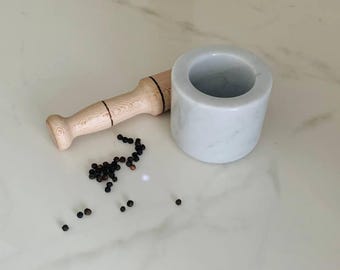 May include: A white marble mortar and pestle set with a wooden handle. Black peppercorns are scattered on the surface. The mortar is cylindrical, and the pestle is light-colored wood with a rounded end.