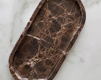 May include: An oval-shaped tray crafted from dark brown marble with prominent white veining throughout the surface. The tray features a smooth, polished finish and a raised rim, showcasing the natural stone patterns and deep earthy color tones.