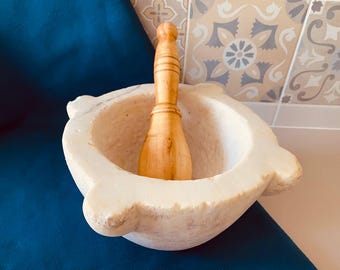 May include: A white marble mortar and pestle set with a wooden pestle. The mortar has a rounded shape with two side handles. The pestle is light brown and fits inside the mortar. The set is on a blue surface.