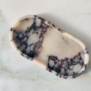 Könnte beinhalten: Oval geformtes Dekotablett mit Marmordesign. Das Tablett hat eine weiße Basis mit dunkelvioletten und grauen Adern. Die Kanten sind glatt und abgerundet. Es ist für kleine Gegenstände wie Schmuck oder Schlüssel gedacht.