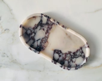 May include: Oval-shaped decorative tray with a marble design. The tray features a white base with dark purple and gray veining throughout. The edges are smooth and rounded. The tray is designed for holding small items.