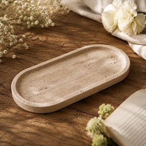 May include: An oval-shaped tray made of beige travertine stone with a porous, textured surface and a raised rim. The tray features natural variations in colour and small indentations characteristic of the stone, presenting a flat, elongated form.