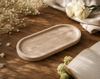 May include: An oval-shaped tray made of beige travertine stone with a porous, textured surface and a raised rim. The tray features natural variations in color and small indentations characteristic of the stone, presenting a flat, elongated form.