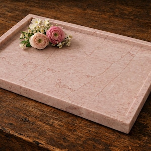 May include: A rectangular pink marble tray with a smooth, polished surface featuring natural veining patterns. The tray has raised edges and is decorated with a small cluster of fresh pink and white ranunculus flowers and baby's breath.