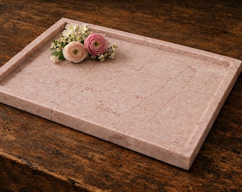 May include: A rectangular pink marble tray with a smooth, polished surface featuring natural veining patterns. The tray has raised edges and is decorated with a small cluster of fresh pink and white ranunculus flowers and baby's breath.