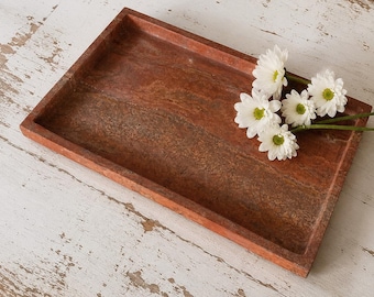 May include: A rectangular, reddish-brown stone tray with a textured, porous surface and raised edges. Resting inside the tray are four white daisy-like flowers with yellow centers and green stems, arranged in a small cluster on the right side.