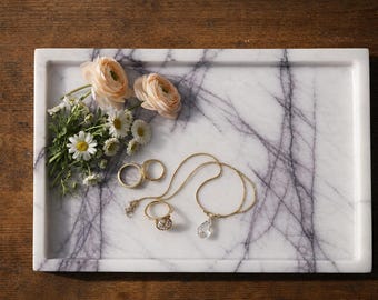 May include: A rectangular white marble tray with purple veining featuring a small bouquet of peach ranunculus and white daisies, two gold bands, a gold ring with a clear stone, and a gold chain necklace with a clear teardrop pendant.
