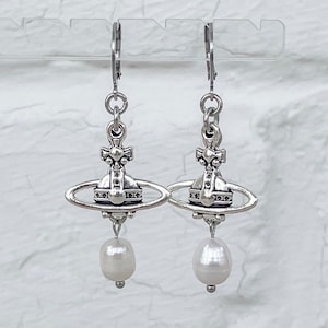 Könnte beinhalten: Silberne Ohrringe mit einer Kugel und einem Perlenanhänger. Die Kugel hat ein detailliertes Design mit einem Kreuz und einem Kreis.