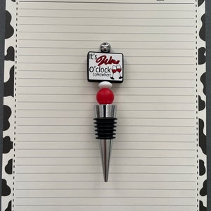 May include: A white and red wine bottle stopper with the text "It's Wine O'clock Somewhere". The stopper has a silver top and a red and white bead.
